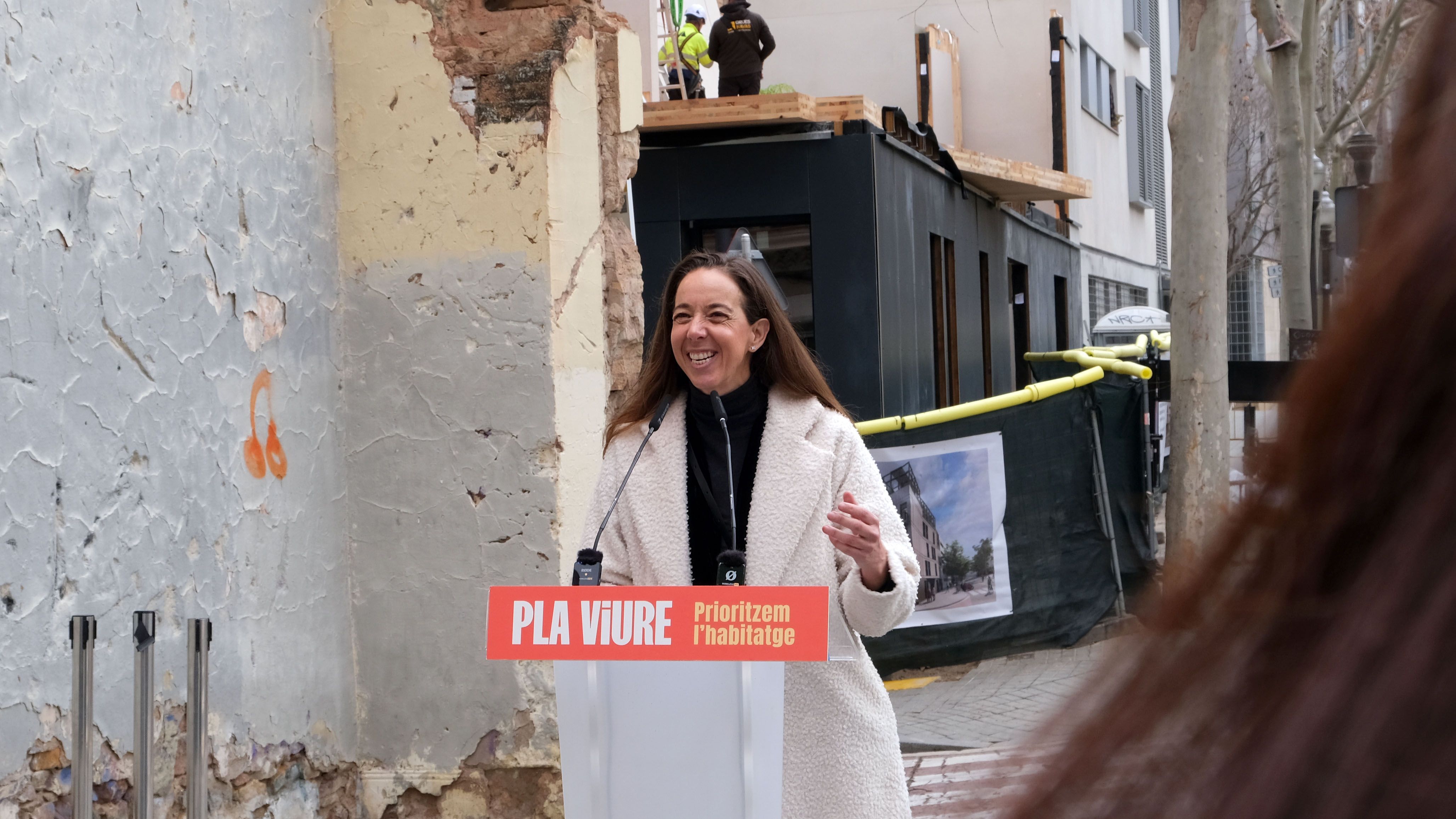 Carlota Pi intervenint en un acte públic davant d’un edifici en rehabilitació, amb el lema “Pla Viure – Prioritzem l’habitatge”.