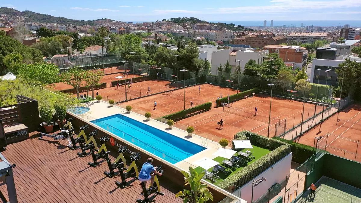 Vista aérea de un complejo deportivo con varias pistas de tenis de tierra batida, piscina y zona de ejercicio al aire libre, con la ciudad y el mar al fondo.