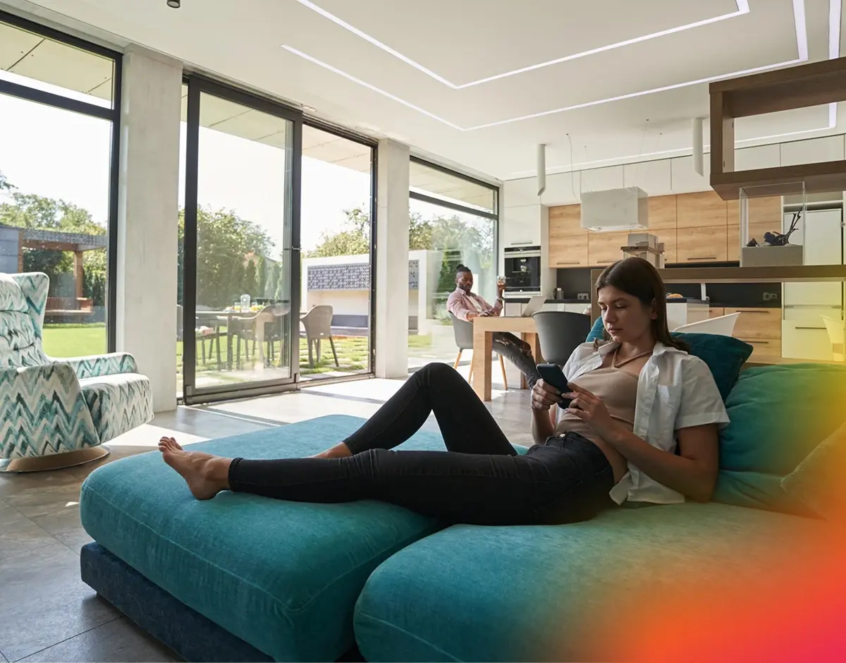 Mujer recostada en un sofá utilizando el teléfono móvil en un salón con grandes ventanales y cocina integrada.